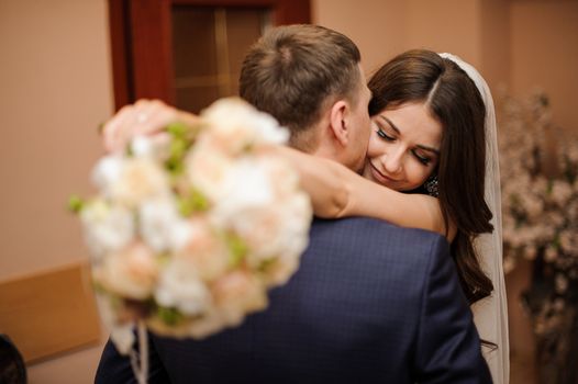 elegant brunette bride with a bouquet of roses hugs a stylish bridegroom, while he kisses her